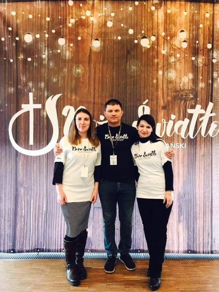 Grupa trzech osób pozuje na tle drewnianej ściany z napisem i dekoracyjnymi lampkami. Wszyscy mają na sobie ubrania z logo 'Boże Światło'.