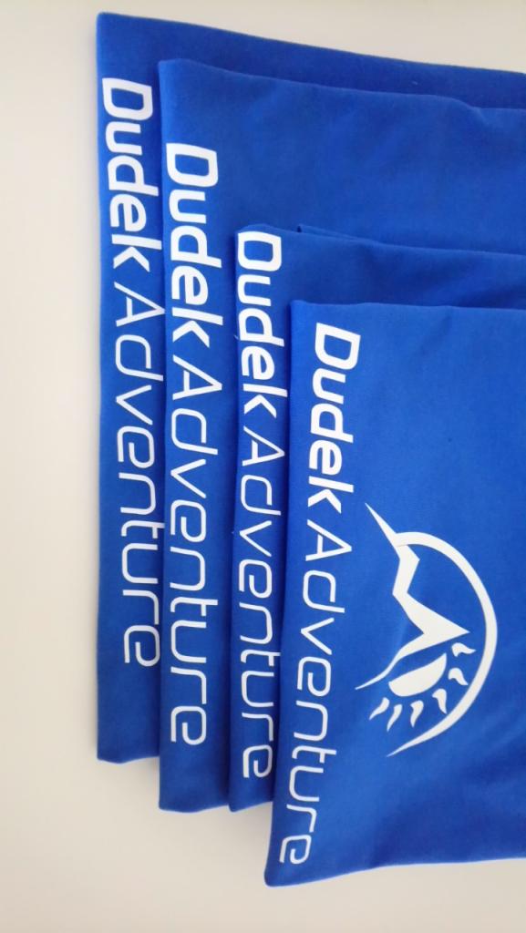 Stos niebieskich koszulek z białym nadrukiem 'Dudek Adventure' i logo, ułożonych pionowo na jasnym tle, widoczne z bliska.