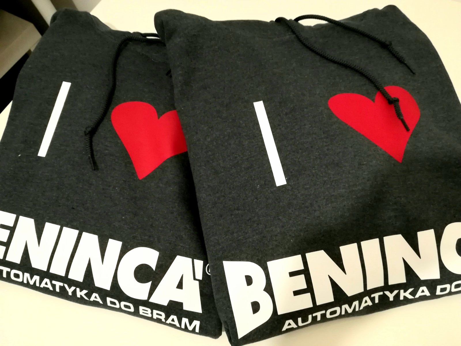 Dwie szare bluzy z kapturem leżące obok siebie, z nadrukiem 'I ❤️ BENING AUTOMATYKA DO BRAM' w kolorze białym i czerwonym.