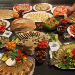 Bogato zastawiony st&oacute;ł z r&oacute;żnorodnymi potrawami, w tym rybą w galarecie z dekoracjami kwiatowymi, pieczywem, sałatkami, jajkami faszerowanymi i pomidorami koktajlowymi, z ręką sięgającą po kanapkę...