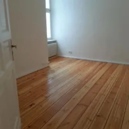 Jasne, drewniane deski podłogowe z widocznymi słojami, świeżo polakierowane, odbijające światło w pustym pokoju z białymi ścianami i otwartymi białymi drzwiami po lewej stronie.