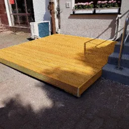 Nowoczesny, niski taras z jasnego drewna, przylegający do budynku z klimatyzatorem i schodami z metalową barierką, położony na brukowanym podłożu.