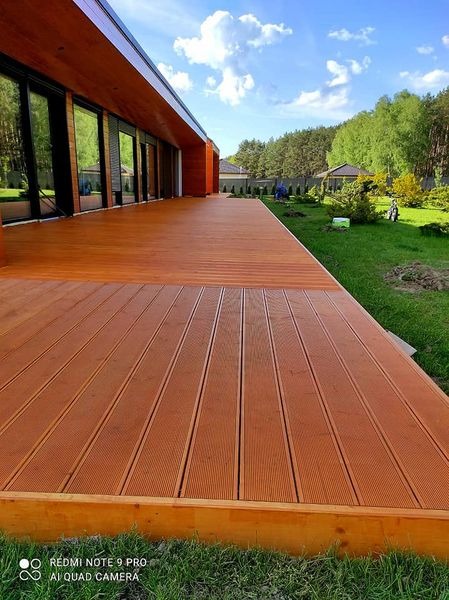 Długi taras kompozytowy w kolorze ciepłego brązu przylegający do nowoczesnego domu z dużymi oknami, widok na zadbany trawnik i drzewa w tle.
