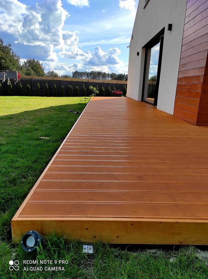 Nowoczesny taras z desek w odcieniach brązu, przylegający do domu z białymi ścianami i drewnianą elewacją, oświetlony lampami wbudowanymi w podest, z widokiem na zielony trawnik i ogrodzenie.