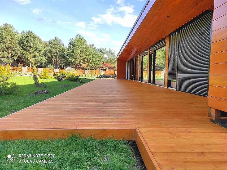 Rozległy, świeżo wykończony taras z desek o ciepłym, miodowym odcieniu, przylegający do nowoczesnego domu z drewnianą elewacją i dużymi oknami z roletami zewnętrznymi, otoczony zielonym trawnikiem...