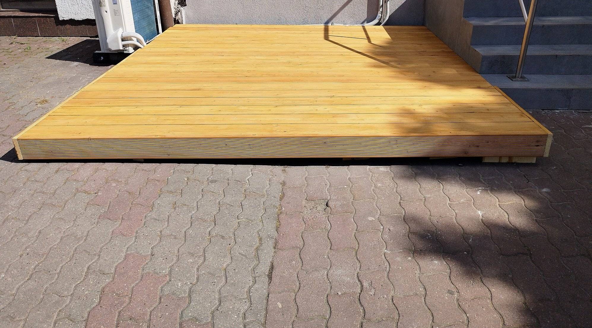 Świeżo wybudowany, prostokątny taras z jasnego drewna, przylegający do budynku z widocznym klimatyzatorem i schodami, ułożony na kostce brukowej w cieniu drzew.