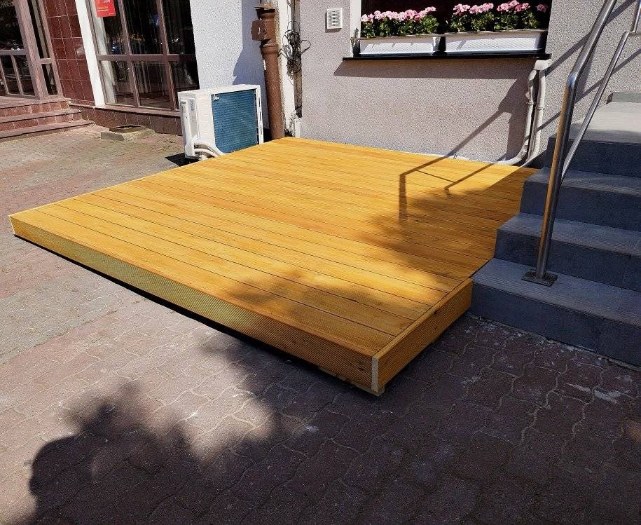 Nowoczesny, niski taras z jasnego drewna, przylegający do budynku z klimatyzatorem i schodami z metalową barierką, położony na brukowanym podłożu.