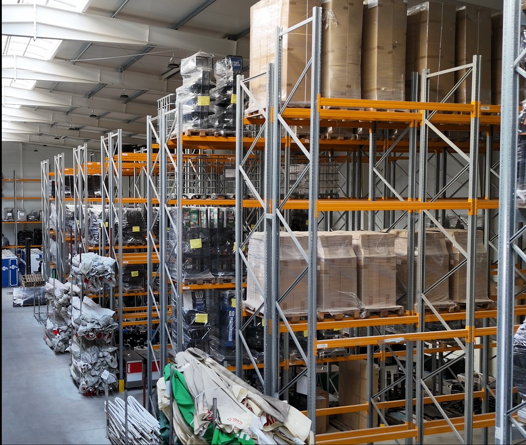 Wnętrze magazynu z wysokimi regałami wypełnionymi zapakowanymi towarami, widoczne palety i metalowa konstrukcja regałów w kolorze srebrnym i pomarańczowym.