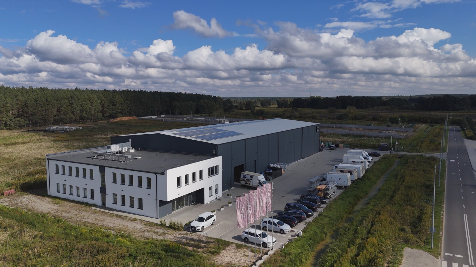 Nowoczesny budynek magazynowy z panelami słonecznymi, widok z lotu ptaka. Zaparkowane busy i samochody osobowe przed budynkiem. Otoczenie: łąka i las.