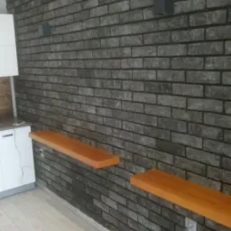 Szara ściana z cegły klinkierowej wewnątrz pomieszczenia, z drewnianymi półkami i minimalistycznym oświetleniem punktowym, widoczny fragment białej szafki i jasna podłoga.