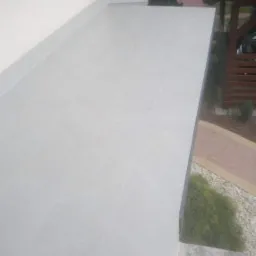 Jasnoszara, płytkowana powierzchnia tarasu przylegająca do białej ściany budynku, widok z góry, z elementami ogrodu w tle.