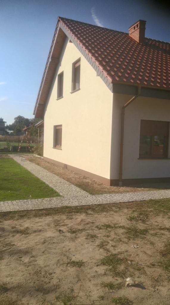 Świeżo otynkowany dom jednorodzinny z czerwoną dachówką, widoczny z boku, z fragmentem chodnika z kostki brukowej i trawnika.