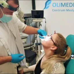Lekarz w masce i rękawiczkach bada nos pacjentki przy użyciu wziernika, z widocznym logo 'Olimedica Centrum Medyczne' w tle.