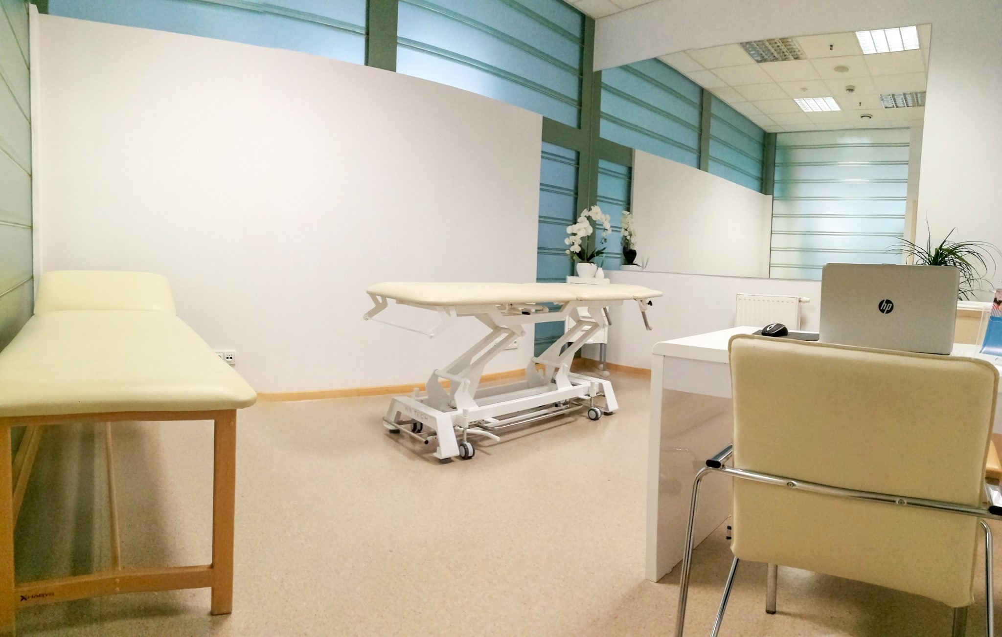Jasne wnętrze gabinetu z dwoma leżankami, biurkiem z laptopem HP i kwiatami, widoczne elementy wyposażenia medycznego.