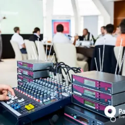 Ręka obsługuje mikser dźwięku podczas konferencji, w tle prezenter i uczestnicy.