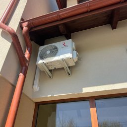 PIOTR MAŁEK ICE SERVICE - Biały klimatyzator Rotenso zamontowany na elewacji budynku z brązowymi rynnami i oknem. Widok z dołu, oświetlenie słoneczne.