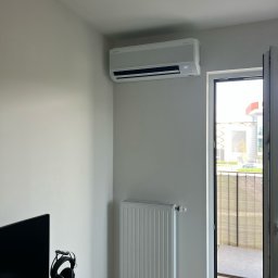 PIOTR MAŁEK ICE SERVICE - Biały klimatyzator na ścianie w pokoju z biurkiem, grzejnikiem i otwartym balkonem. Widok na budynek zza balkonu. Nowoczesny design, jasne wnętrze.