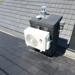 PIOTR MAŁEK ICE SERVICE - Biały klimatyzator marki Gree zamontowany na czarnym kominie na dachu z grafitowej dachówki. Widoczne przewody i elementy instalacji. Słoneczny dzień.