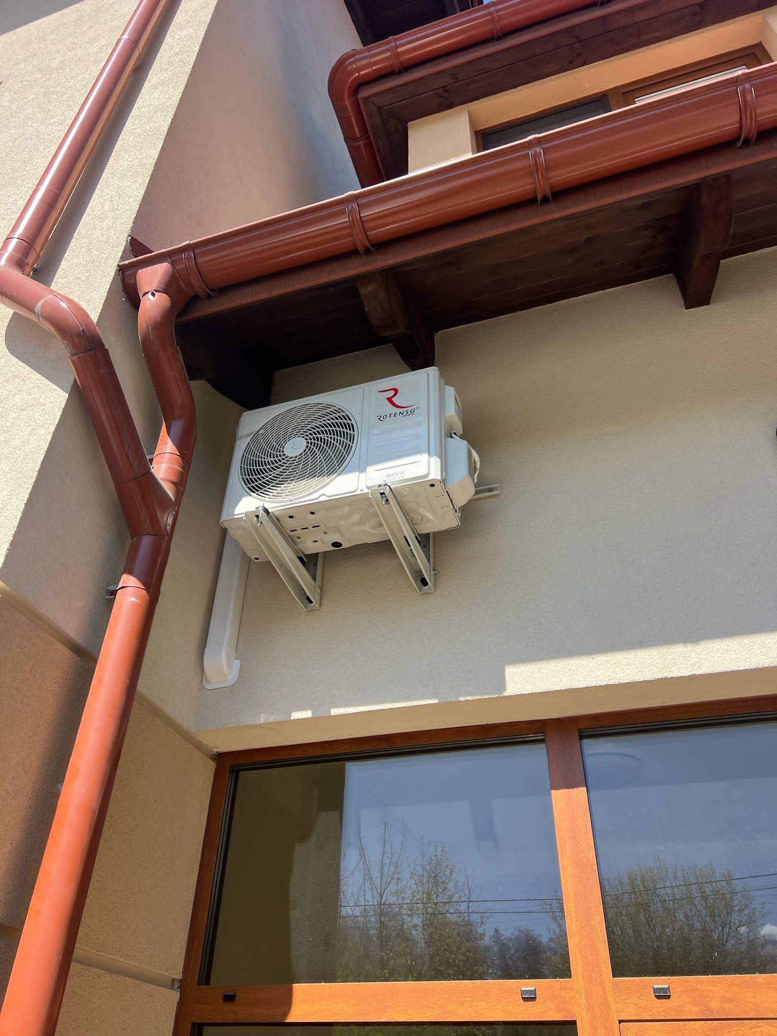 Biały klimatyzator Rotenso zamontowany na elewacji budynku z brązowymi rynnami i oknem. Widok z dołu, oświetlenie słoneczne.