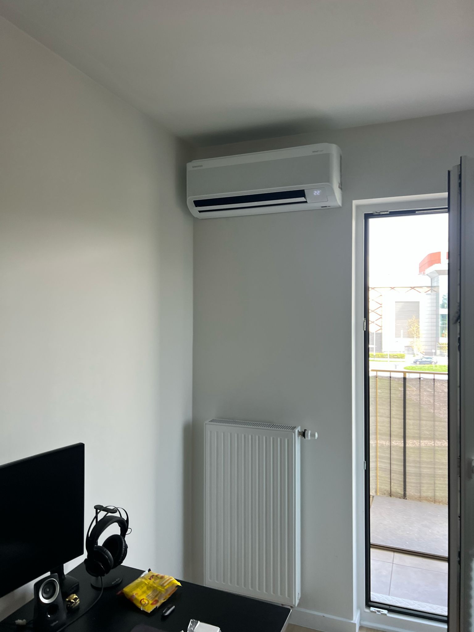 Biały klimatyzator na ścianie w pokoju z biurkiem, grzejnikiem i otwartym balkonem. Widok na budynek zza balkonu. Nowoczesny design, jasne wnętrze.