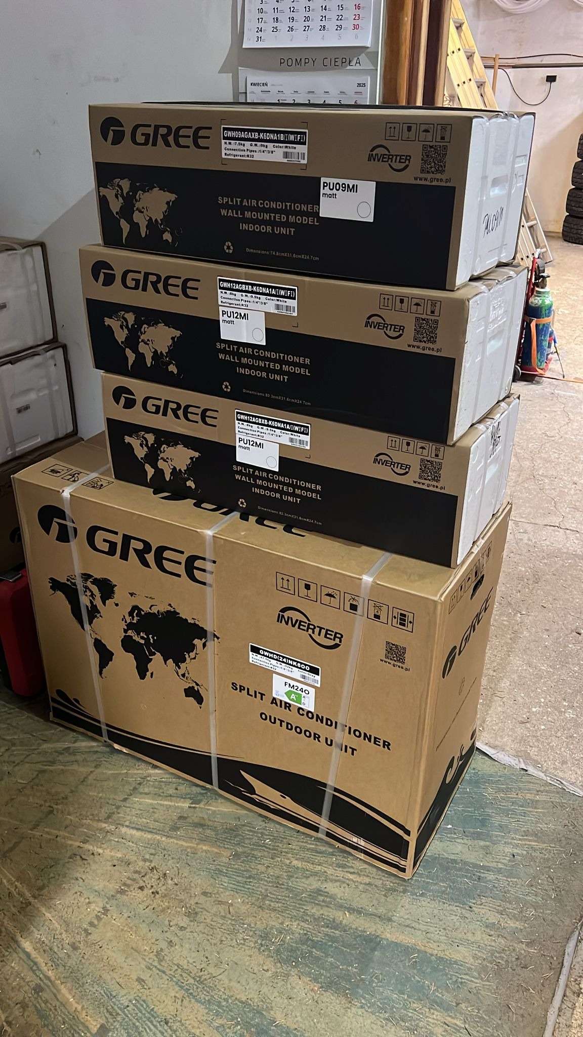 Stos kartonów klimatyzatorów Gree, modele ścienne i zewnętrzne, z widocznymi oznaczeniami i specyfikacjami technicznymi. Tło magazynowe, widoczny kalendarz.