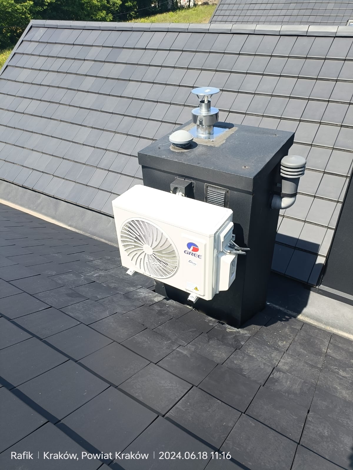 Biały klimatyzator marki Gree zamontowany na czarnym kominie na dachu z grafitowej dachówki. Widoczne przewody i elementy instalacji. Słoneczny dzień.