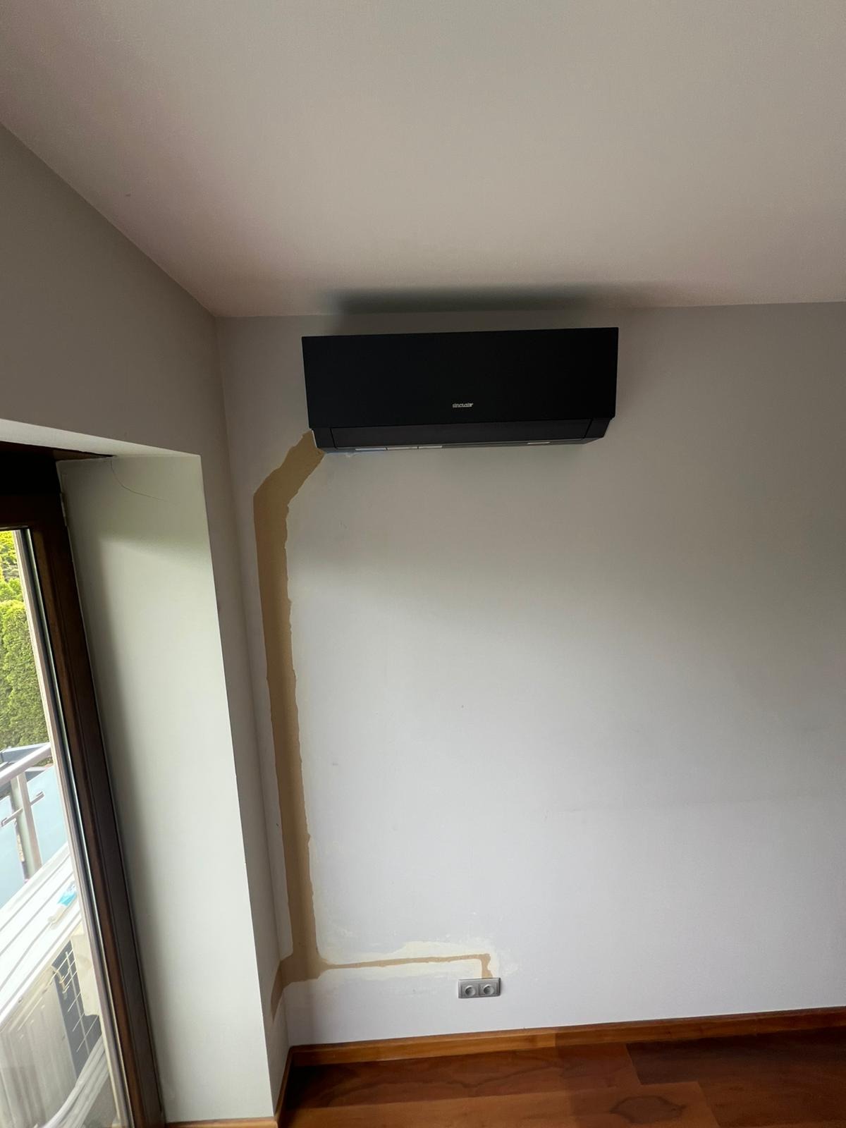 Czarny klimatyzator Sinclair na ścianie, widoczne ślady po instalacji przewodów. Podłoga z paneli, okno z widokiem na zieleń. Ujęcie z dołu, podkreślające montaż.
