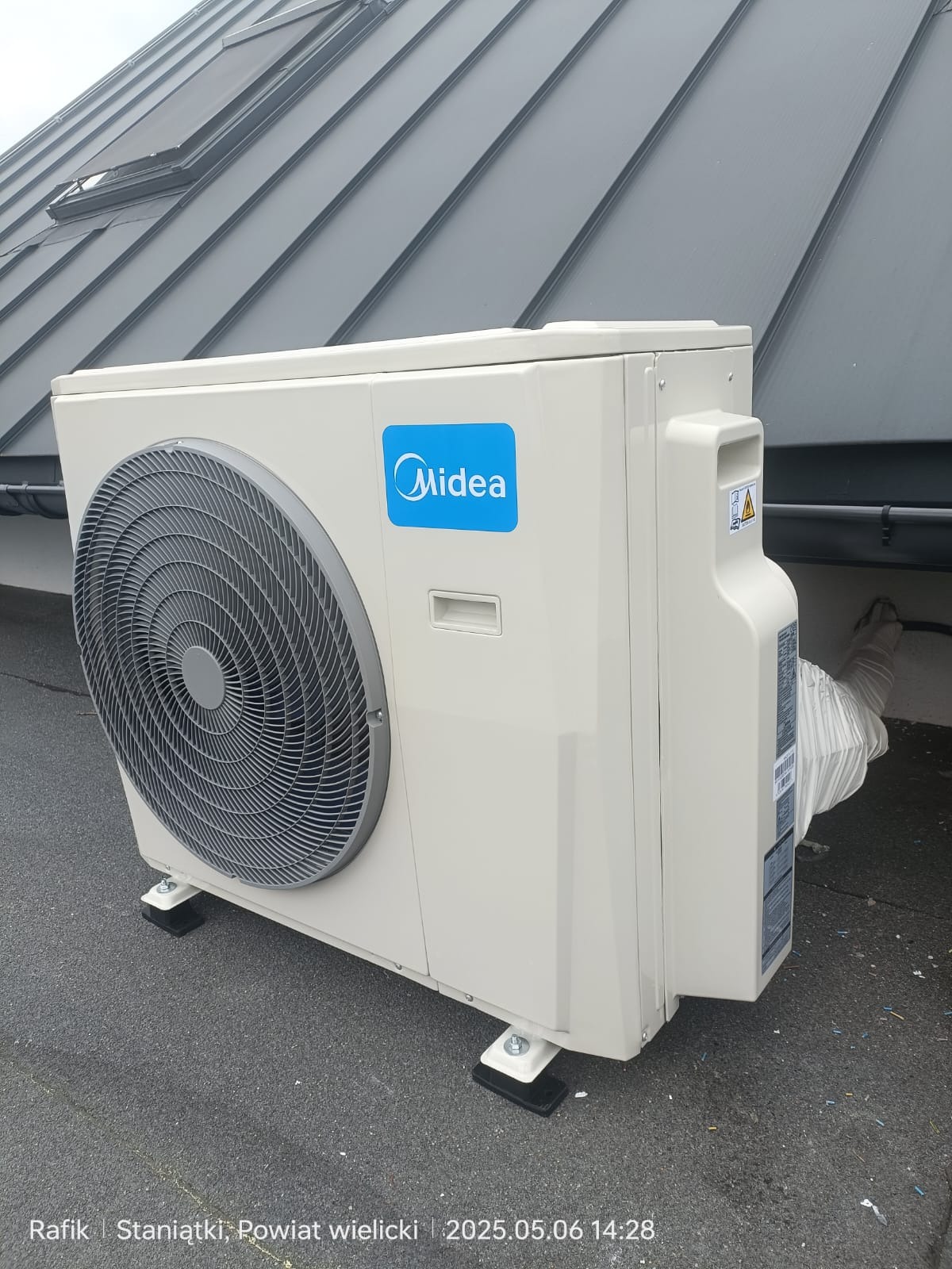 Zewnętrzna jednostka klimatyzacji Midea na dachu z blachy, widoczny fragment rynny i okno dachowe. Urządzenie stoi na gumowych podkładkach antywibracyjnych.