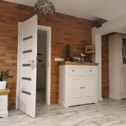 Ceglana ściana w salonie z otwartymi białymi drzwiami z poziomymi przeszkleniami, białe meble w stylu retro, kwiat w doniczce i lampa Tiffany