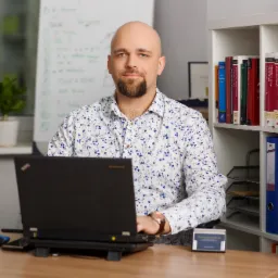 Portret mężczyzny z brodą w kwiecistej koszuli siedzącego za biurkiem z laptopem, obok statuetka Temidy i regał z książkami prawniczymi, w tle tablica suchościeralna z notatkami.