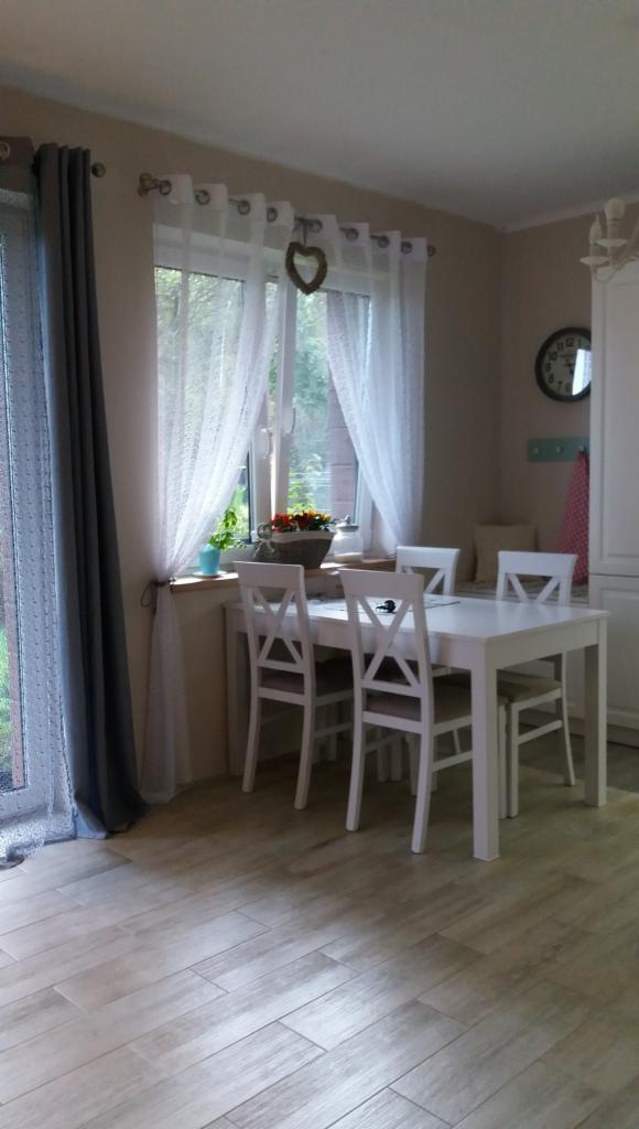 Jasna jadalnia z białym stołem i krzesłami, drewnopodobne płytki na podłodze, okno z koronkowymi firankami i szarymi zasłonami.