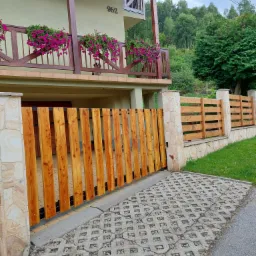 Nowoczesna drewniana brama wjazdowa z pionowych desek, osadzona w kamiennych słupkach, prowadząca na podjazd z ażurowych płyt betonowych, z widokiem na piętrowy dom z balkonem i kwiatami.