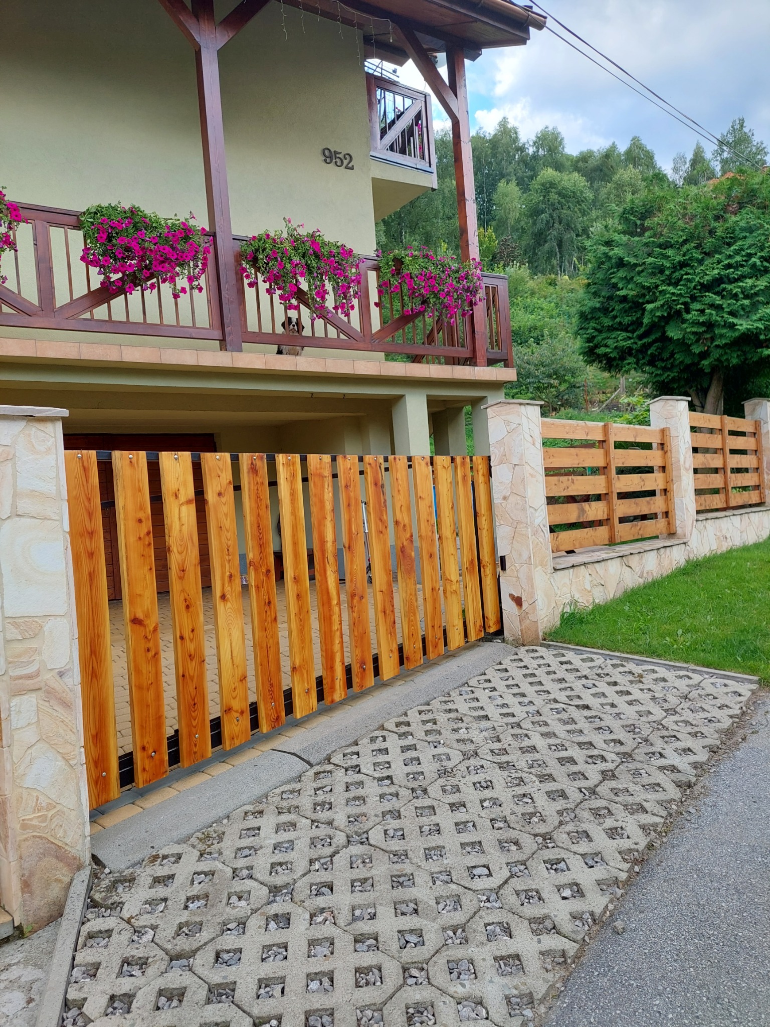 Nowoczesna drewniana brama wjazdowa z pionowych desek, osadzona w kamiennych słupkach, prowadząca na podjazd z ażurowych płyt betonowych, z widokiem na piętrowy dom z balkonem i kwiatami.