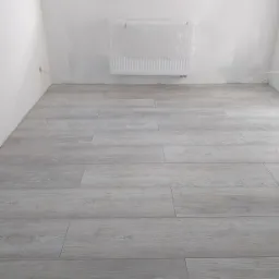 Nowo ułożone panele podłogowe w jasnoszarym odcieniu, imitujące drewno, w pustym, jasnym pokoju z grzejnikiem zabezpieczonym folią malarską i białymi ścianami.