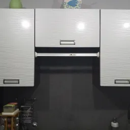 Białe szafki kuchenne wiszące z poziomymi, falistymi wzorami na frontach, srebrne uchwyty, ciemnoszara ściana w tle, widoczny fragment okapu i zlewu z baterią.