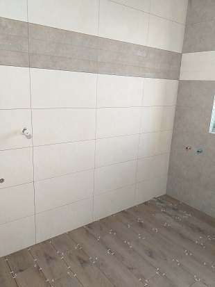 Wnętrze łazienki w trakcie układania płytek: białe płytki ścienne z szarym pasem dekoracyjnym, podłoga z paneli imitujących drewno z widocznymi krzyżykami dystansowymi.