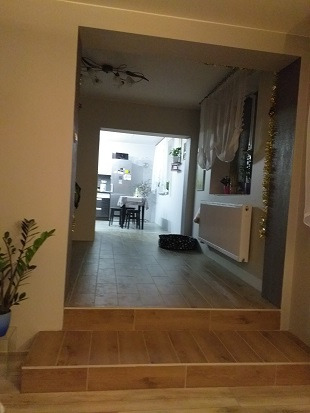 Widok z salonu na kuchnię przez podwyższone przejście z dwustopniowymi schodkami wyłożonymi płytkami imitującymi drewno, w tle widoczny stół i krzesła w kuchni, a na ścianie grzejnik i dekoracje.