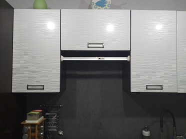 Białe szafki kuchenne wiszące z poziomymi, falistymi wzorami na frontach, srebrne uchwyty, ciemnoszara ściana w tle, widoczny fragment okapu i zlewu z baterią.