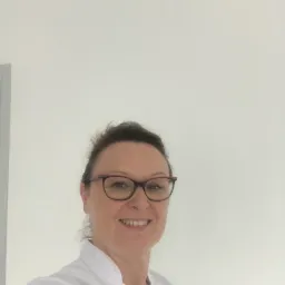 Uśmiechnięta kobieta w okularach i białym uniformie, prawdopodobnie pracownik służby zdrowia, na tle jasnej ściany.