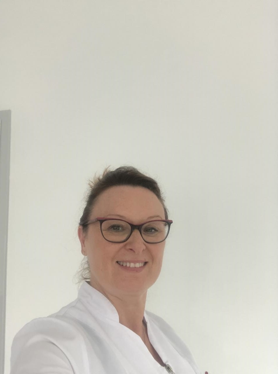 Uśmiechnięta kobieta w okularach i białym uniformie, prawdopodobnie pracownik służby zdrowia, na tle jasnej ściany.