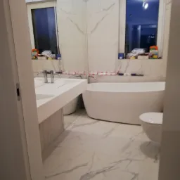 Nowoczesna łazienka z wanną wolnostojącą, umywalką nablatową, toaletą i płytkami imitującymi marmur na podłodze i ścianach, widok zza otwartych drzwi.