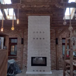 Biały, kaflowy piec kominkowy z czarnym wkładem, w rustykalnym wnętrzu z drewnianym stropem, ceglanymi ścianami i wiszącymi żarówkami na sznurkach.