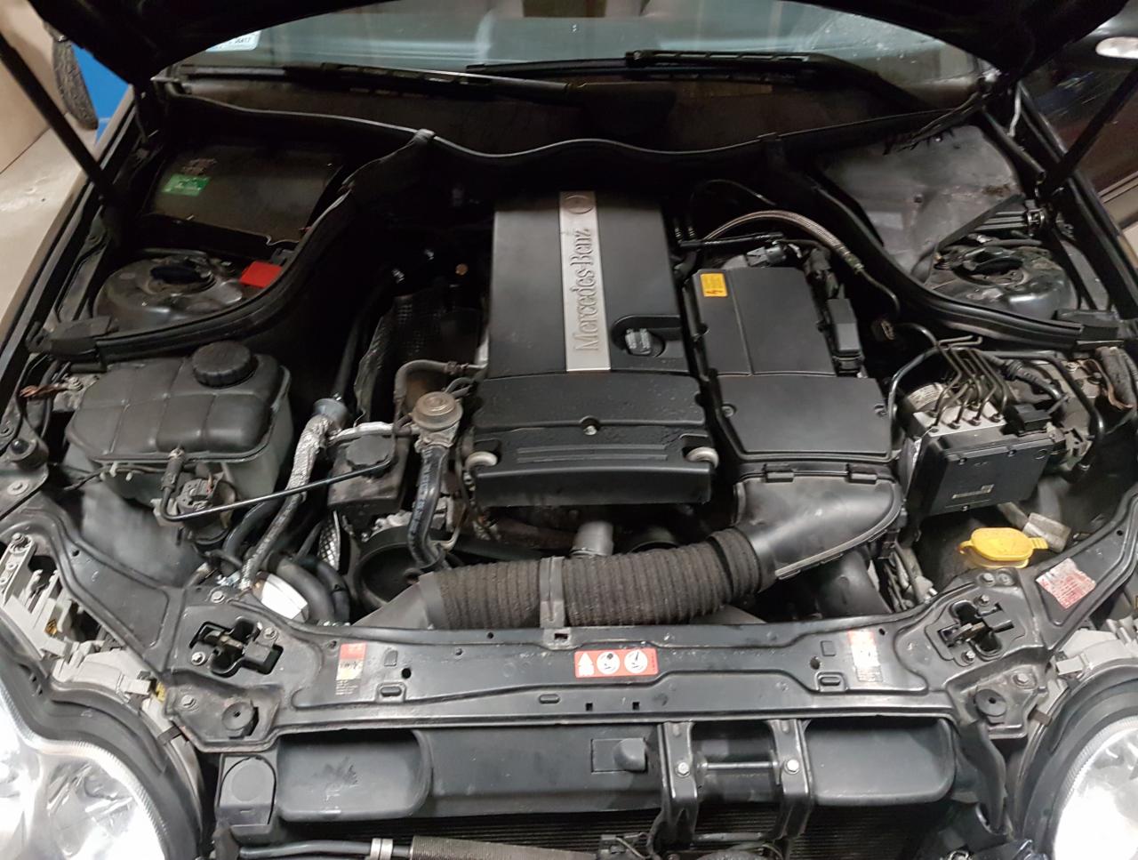 Otwarta maska samochodu Mercedes-Benz, widoczny silnik z elementami instalacji paliwowej, czarna obudowa silnika z napisem Mercedes-Benz.