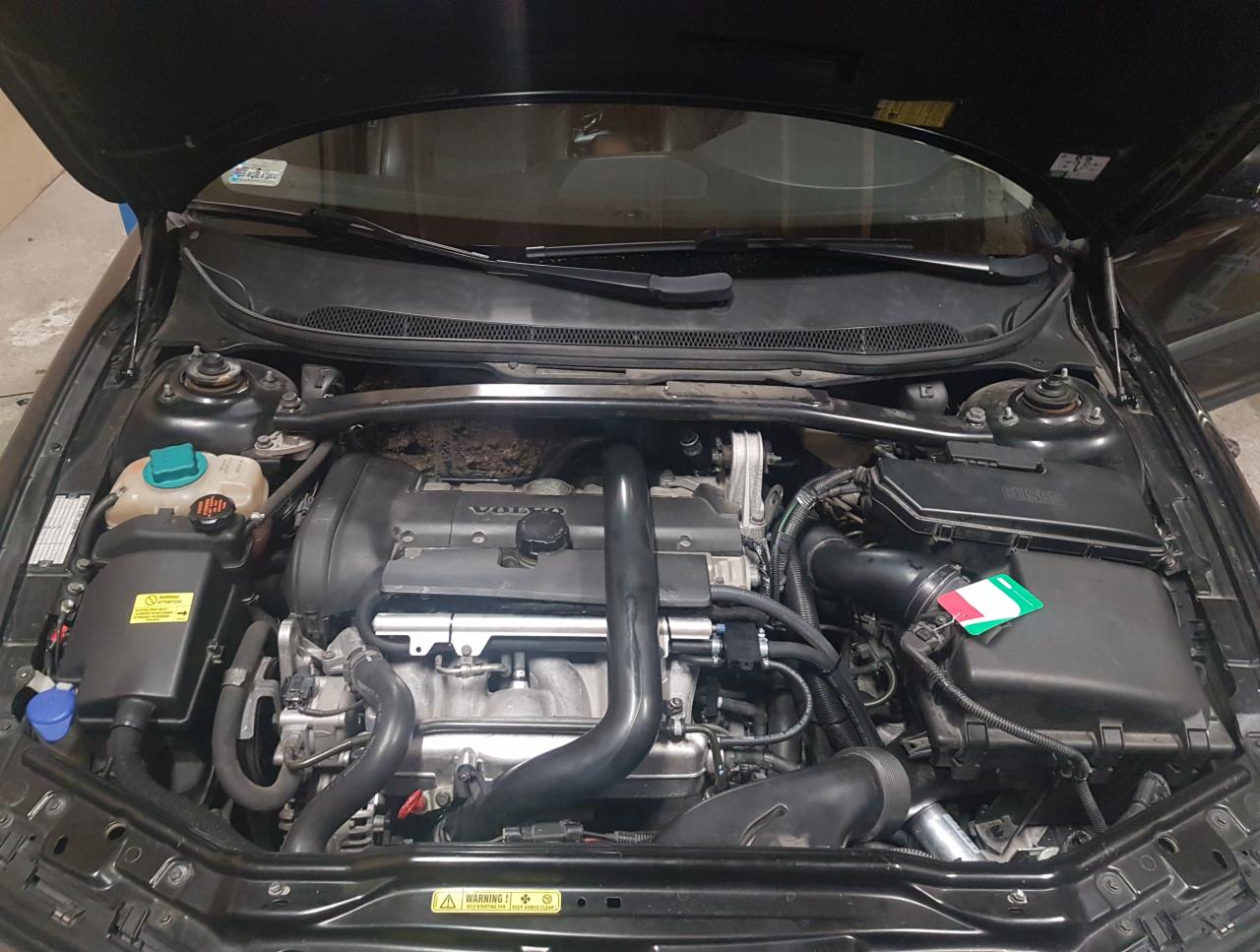 Otwarta maska czarnego samochodu marki Volvo, widoczny silnik z elementami instalacji zasilania paliwem alternatywnym, na obudowie filtra powietrza naklejka z flagą Włoch.