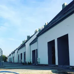 Szereg identycznych domów jednorodzinnych z białymi, świeżo otynkowanymi elewacjami i grafitowymi wnękami wejściowymi, widok z perspektywy chodnika z kostki brukowej.