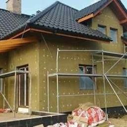 Usługi Ogólnobudowlane Dawid Plesner - Dom w Wołowie w trakcie ocieplania elewacji, widoczne rusztowania i płyty izolacyjne mocowane do ściany. Częściowo ocieplona ściana w kolorze beżowym, widoczne punkty mocowania. Na pierwszym planie...