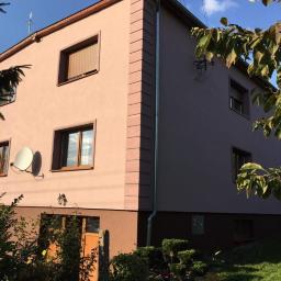 Usługi Ogólnobudowlane Dawid Plesner - Dwukondygnacyjny dom z ocieploną elewacją w odcieniach beżu i brązu, z widocznymi detalami architektonicznymi w postaci boniowania narożników, brązowych ram okiennych i rolet zewnętrznych, antena...