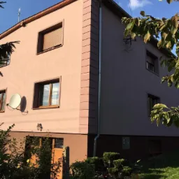 Dwukondygnacyjny dom z ocieploną elewacją w odcieniach beżu i brązu, z widocznymi detalami architektonicznymi w postaci boniowania narożników, brązowych ram okiennych i rolet zewnętrznych, antena...