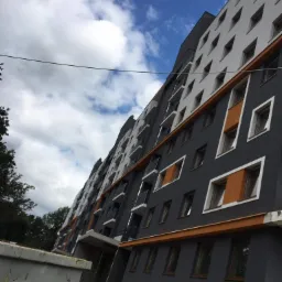 Nowoczesny budynek wielorodzinny z szarą elewacją, białymi balkonami i pomarańczowymi akcentami wokół okien, sfotografowany pod kątem z dołu, z widocznym fragmentem kontenera budowlanego.