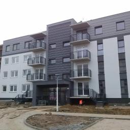 Usługi Ogólnobudowlane Dawid Plesner - Nowoczesny budynek wielorodzinny z balkonami w odcieniach szarości i bieli, teren wokół w trakcie prac wykończeniowych, widoczny pracownik w pomarańczowej kamizelce.